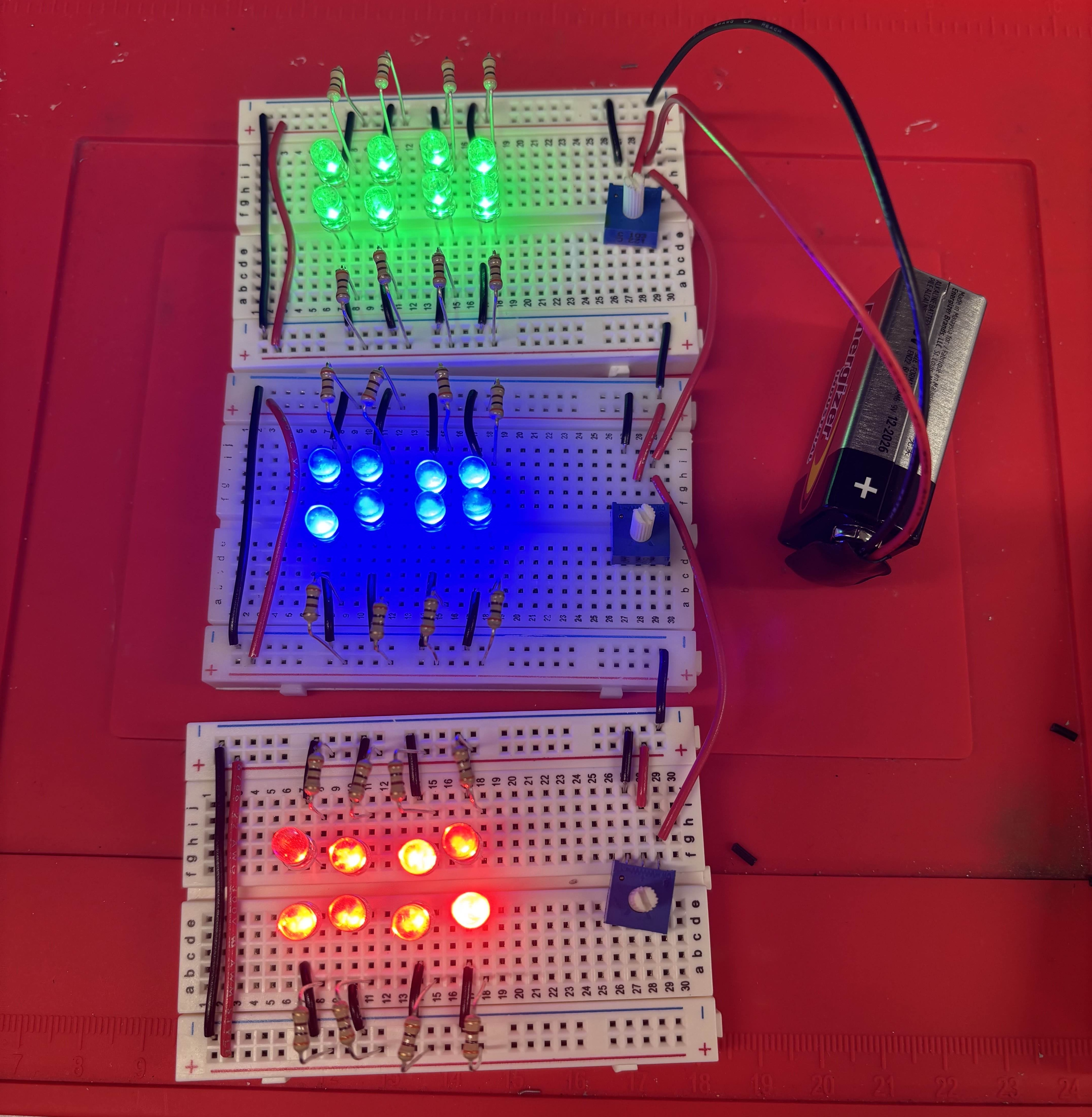 Three breadboards stacked, each with a different coloured LED lit up, all connected to a 9V power source.